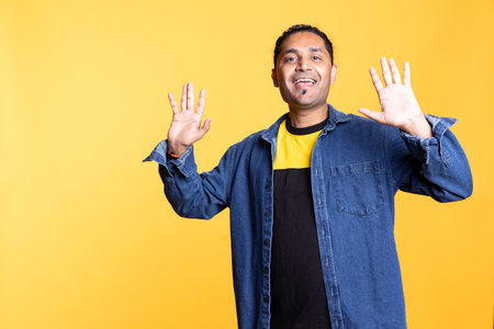 Indian friendly male person with a man bun raising his hands and waving, greeting his friends against orange background. Young adult in a casual clothes, joyous satisfied guy.の写真素材