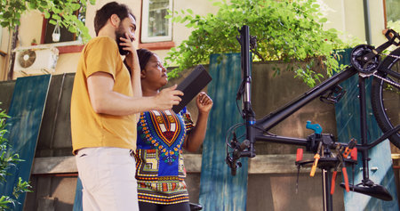 Youthful multiethnic couple utilizing digital device for repairing and maintaining modern bicycle. In home yard vibrant man and sporty black woman exploring the internet for bike maintenance.の写真素材