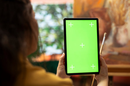 Painter holding a device with isolated mockup screen in her art studio, looking at her finished masterpiece with oil paints on canvas. Beautiful rustic artwork for a fine art gallery, creativity.の写真素材