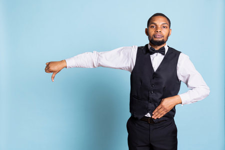 Displeased negative bellboy gives a thumbs down sign in studio, showing rejection and disapproval. Dissatisfied discontent hotel personnel with tuxedo does dislike rejection gesture.の写真素材