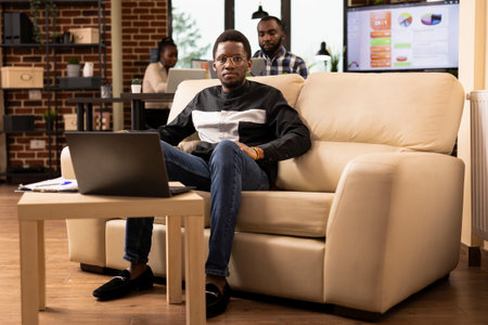 Portrait of african american businessman with glasses, sitting on cozy sofa and looking at camera. Black male employee with laptop on table, seated at lounge area in brick wall office, ready for work.の写真素材