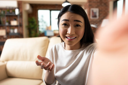 Joyful self employed woman gesturing to camera during home livestream, sharing business insights. Confident asian woman connects with online audience through friendly video content.の写真素材