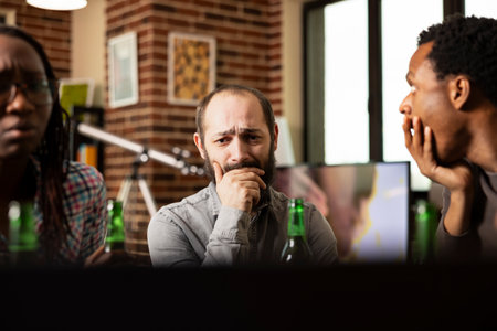 Concerned white man watching emotional content with african american friends in brick wall room. Bearded male individual sitting at table, appears distressed while viewing sad video with covered mouthの写真素材