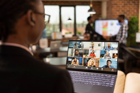 Rearview of female business advisor in videoconference with remote coworkers, laptop screen displaying the virtual meeting. African American professional on video call, attending online discussion.の写真素材