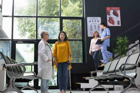 Woman walking with crutches during health check at modern waiting area, receiving support from general practitioner. Medic offering physical support while guiding recovery journey.の写真素材