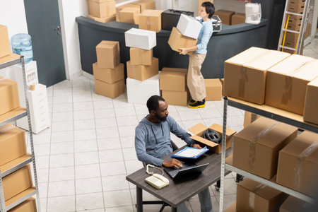 Black warehouse worker handling cargo and shipping logistics, coordinating inventory and hand packing merchandise, industrial warehouse shelves. Express delivery service at desk.の写真素材
