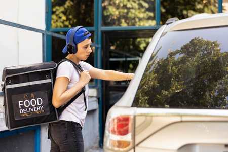 Confident woman delivering takeout with the car using a backpack, arriving at the entrance of a modern building. Employee preparing to ring the door bell to bring the food order to client.の写真素材