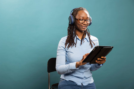 Attractive young black woman with wireless headphones touches her tablet while watching videos. African american lady seated on chair with blue shirt and background, enjoying digital content.の写真素材