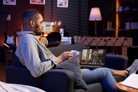 Male remote worker waving at his associates during virtual meeting, showcasing effectiveness of online tools and internet connection. Young adult sharing insight in a briefing session.の写真素材
