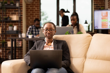 Black female manager sits on cozy couch, using laptop to analyze startup research data and manage company reports. Focused businesswoman with glasses, reviewing emails and preparing business projects.の写真素材