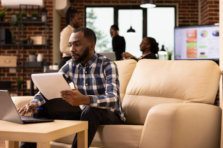 Black male employee updates financial records on his laptop, referencing a clipboard in startup office. Focused businessman reviews company paperwork, using digital tools to manage business reports.の写真素材