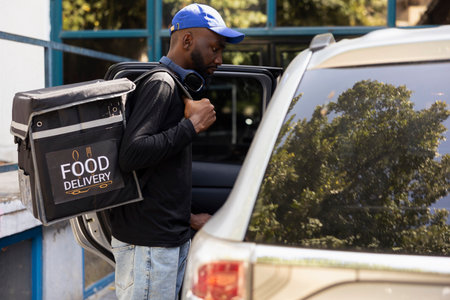 Male courier arriving at drop off address by car and delivering meal, taking out backpack to bring fast food to a client at the front door. Takeaway delivery service from a restaurant.の写真素材