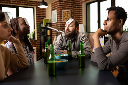 Diverse male and female individuals playing cards and sharing snacks in brick wall room. Young adults sitting around table with drinks and bowl of chips, enjoying friendly indoor game session.の写真素材