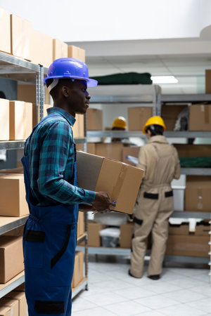 African american worker in protective gear checks merchandise for dispatch in a busy warehouse. Crates and boxes arranged for logistics and transportation in operational supply chain hub.の写真素材