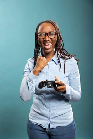 Smiling african american woman holding gamepad, enjoying a fun video game in blue background studio. Happy black female gamer with modern controller, celebrating a victory with fist raised.の写真素材