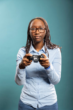 Black woman with glasses holding gaming controller and having fun playing video games in modern studio. African american female model in blue shirt fully engaged in a serious gaming session.の写真素材