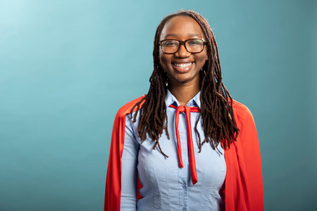 Portrait of black female individual cheerfully standing isolated over blue background. Smiling young lady with glasses and red superhero cape, looking at camera, portraying positivity in modern studioの写真素材
