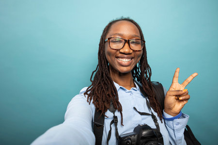 African american female vlogger giving peace sign to her online audience, recording herself in studio. Pov of excited black woman photographer making two finger, v gesture towards the camera.の写真素材
