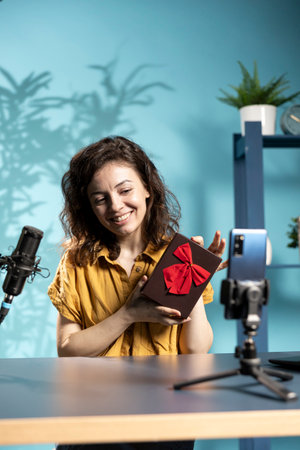 Joyful content creator holding present box, filming giveaway using selfie phone camera. Woman happy to offer packed gift with ribbon to audience, recording with smartphone on selfie stickの写真素材