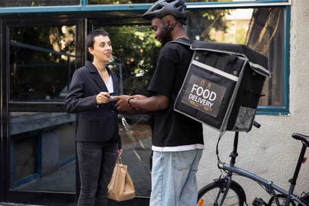 Woman client paying for her takeaway order with a contactless card at the doorstep. Delivery man carrying a thermal bag, completes fast food transaction using a wireless POS terminal.の写真素材