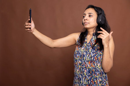 Gorgeous female vlogger wearing wireless headphones, smiling as she holds her smartphone to record...
