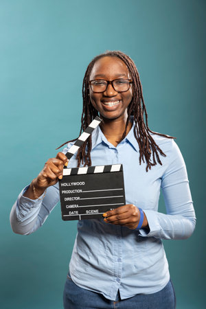Portrait of african american female movie director happily holding clapperboard, ready for shooting. Smiling female filmmaker poses with film slate in hand, looking at camera with enthusiasm.の写真素材