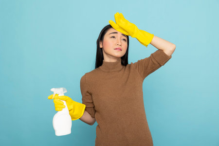 Fatigued female janitor in yellow rubber gloves touches forehead in exhaustion while holding cleaning spray. Overworked asian housekeeper sadly looking up, reflecting stress after chores.の写真素材