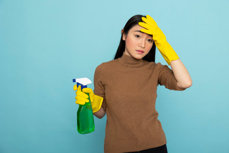 Exhausted female janitor in rubber gloves grasping spray bottle, wiping forehead after intense...