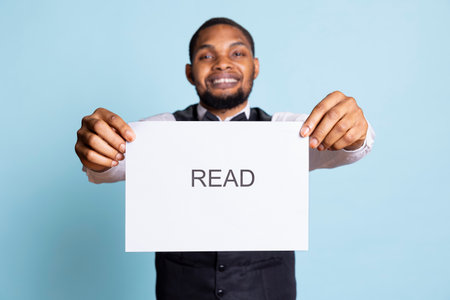 African american hotel personnel getting the attention of clients with an important message, asking them to read the note on the paper sign. Bellboy with professionalism working in tourism.の写真素材