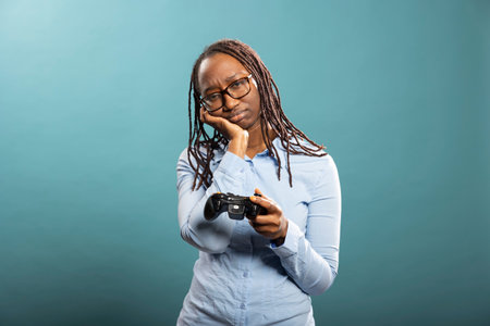 African american woman resting her head on one hand with annoyance as she holds modern controller,...