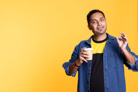 Happy confident indian man serving a coffee from disposable paper cup against orange background, wearing casual clothes and enjoying fresh caffeine refreshment in the morning. Studio beverage.の写真素材