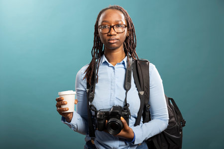 Young woman poses in studio, dressed casually with dslr camera hanging on her neck and coffee in hand. African american female model shows a focused yet professional photographer look.の写真素材