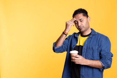 Sleepy man yawning and rubbing eyes sipping coffee to wake up and gain energy. Exhausted person feeling fatigued after sleepless night, drinking caffeinated beverage, orange background.の写真素材