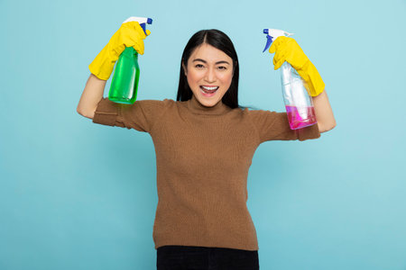 Excited asian housekeeper wearing yellow gloves and flexing arms while holding pink and green...