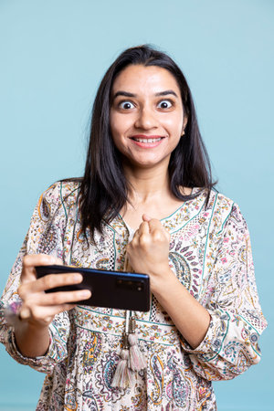Delighted pleased girl winning the mobile video games in studio, celebrating win and achievement against blue background. Young woman feels excited loving to play gaming competition.の写真素材