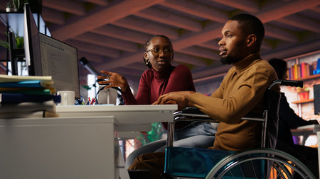 Manager coming to inclusive startup office employee in wheelchair desk to offer help. Thoughtful supervisor checking up on software development agency worker with disability, camera Bの写真素材
