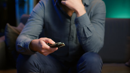 Close up of man in dimly lit apartment using remote control to browse though movies on streaming service. Remote control used by person in living room flipping through channels on TV, camera Aの写真素材