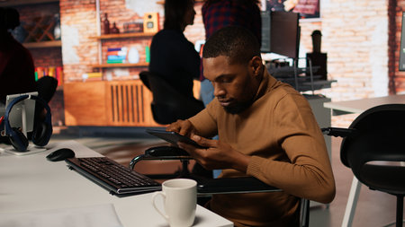 Admin in wheelchair using tablet in innovative startup office to build application. Inclusive software development agency worker with paraplegia checks code on device, camera Aの写真素材