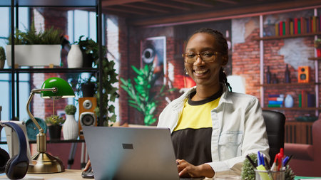 Portrait of cheerful woman in home office opening laptop, starting working on tasks. Joyous african american person using electronic notebook device to do job online, camera Aの写真素材