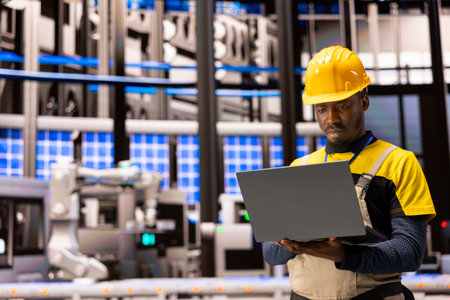 Black engineer monitors industry 4.0 system in industrial plant. Surrounded by robotic arms and...