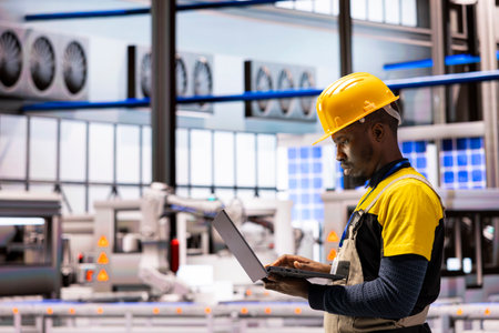 Engineer in smart manufacturing plant checks performance of automated systems on laptop. Integration of robotic arms and high tech machinery showcases Industry 4.0 advancements, optimizing production.の写真素材