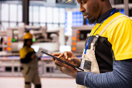 Black male engineer monitors digital automation systems in smart manufacturing facility, optimizing...