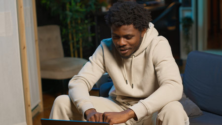 Independent black man using laptop for remote job tasks on a coffee table, surrounded by notes and...