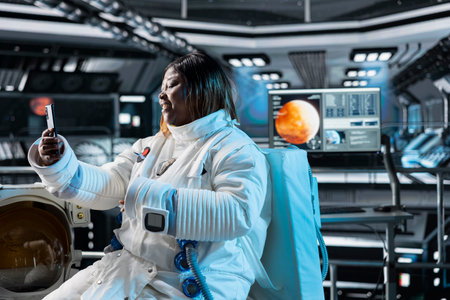 Cheerful cosmonaut in starship using smartphone to do videocall with loved ones back home. Happy...