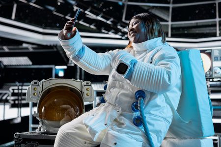 Cheerful female astronaut in starship travelling to space doing selfies using cellphone camera. Happy woman aboard orbit station having fun doing pictures on phone during cosmic journeyの写真素材