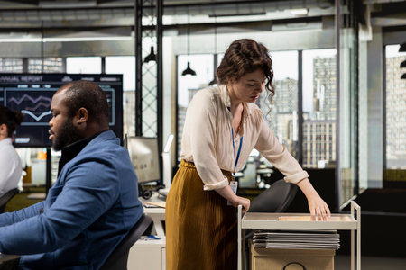 Workplace support employee moves a delivery cart stacked with folders and contracts, helping african american administrator bringing stack of archived documents for accounting tasks.の写真素材