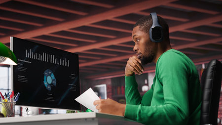 Teleworker in home office watching business conference on PC, looking over statistics charts. African american remote employee in apartment looking at presentation graphs on computer. Camera A.の写真素材