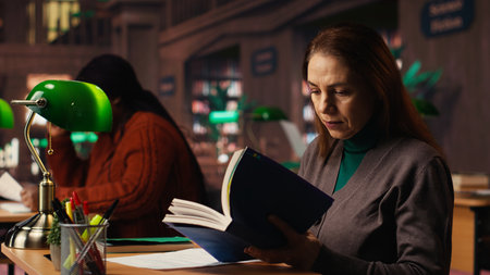 Determined woman writing thesis citations from a specialty book at the library, mature learner working toward a higher education goal. Academic materials in a quiet campus study area.の写真素材