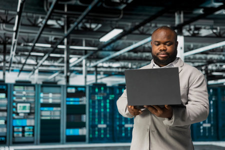 Technician in server hub managing infrastructure systems, checking recovery plan on laptop. Data center employee monitoring performance using tools, making sure rigs are functioning optimallyの写真素材