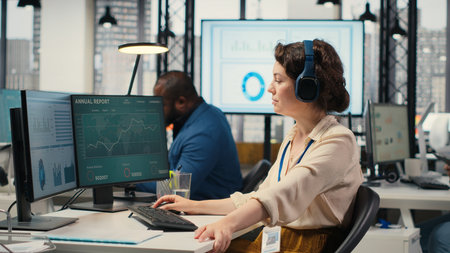 Office administrator uses focus music on headset to solve tasks, monitoring performance metrics at the workstation. Administrative tasks to ensure enterprise productivity and success. Camera B.の写真素材